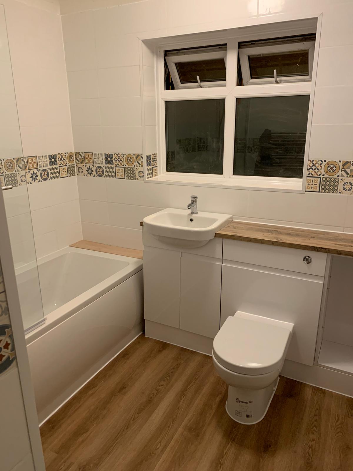A finished bathroom with a bath, vanity basin with oak-effect worktop and patterned border tiles under the window